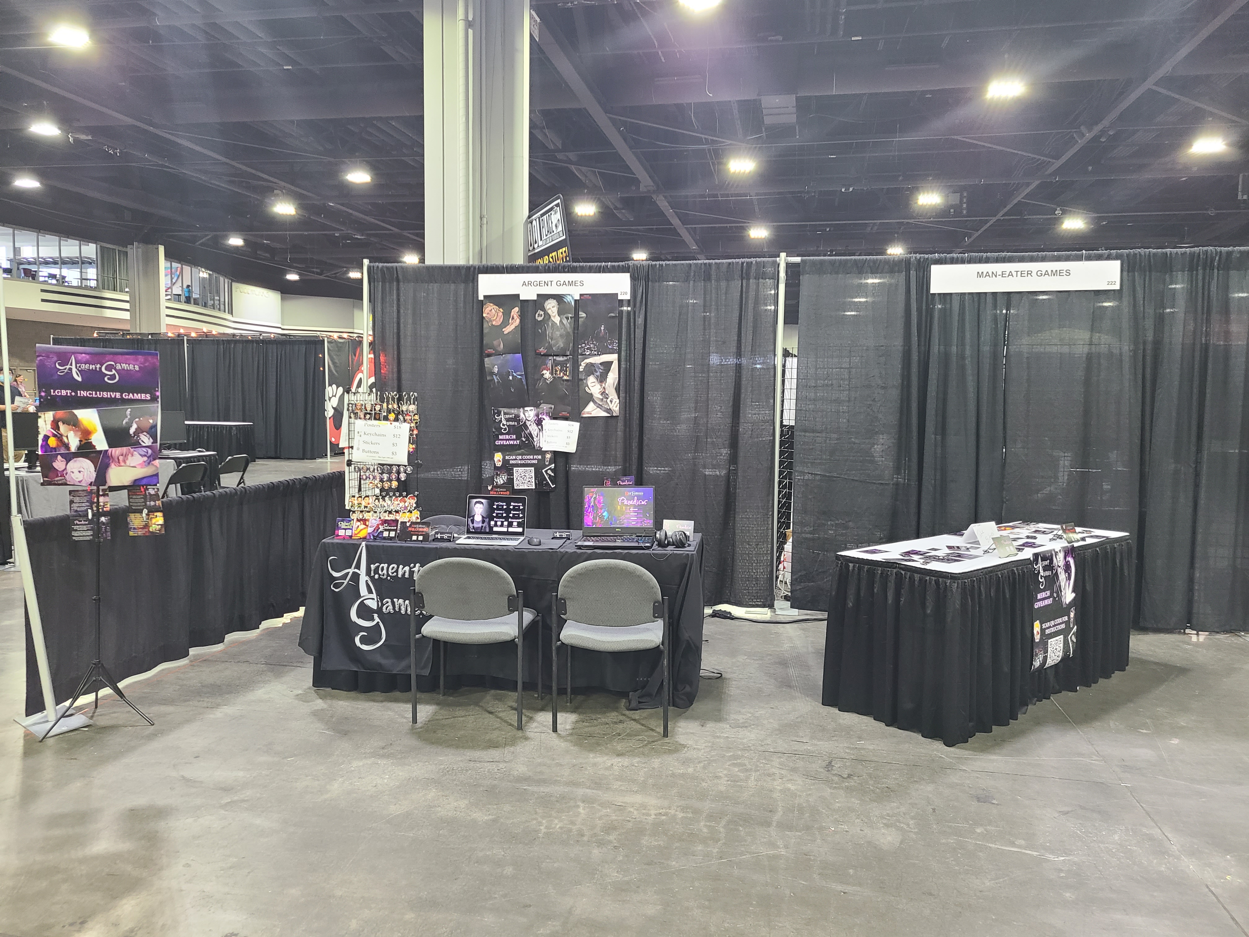 day 3 exhibit booth setup with two tables: one with two computers and the merch stand, and another with cards and buttons spread out on top