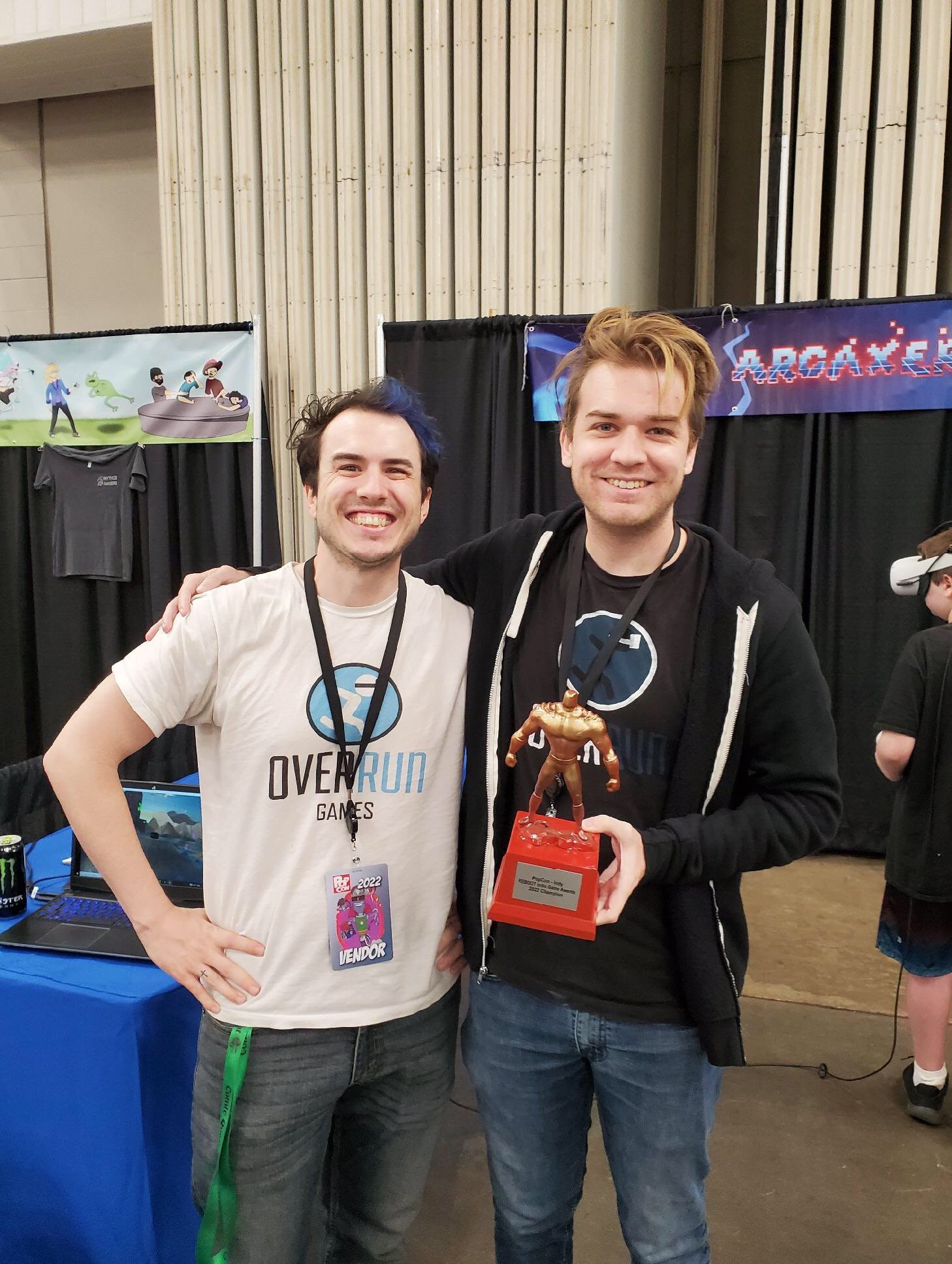 the two arcaxer devs posing with the vr game awards trophy in front  of their booth